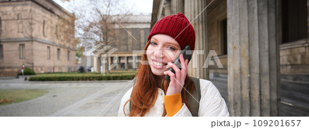 Smiling young redhead woman listens to voice message, makes a phone call, walks on street and talks to someone on smartphone Smiling young redhead woman listens to voice message, makes a phone call, walks on street and talks to someone on smartphone 109201657