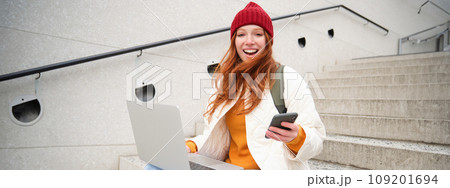 Smiling redhead woman with mobile phone and laptop, sitting on stairs outside building, connects to public wifi, using smartphone and computer 109201694