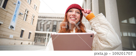 Happy redhead girl in red hat, walks around city with digital tablet, connects to public internet wifi and looks for route, looks at map on her gadget 109201707