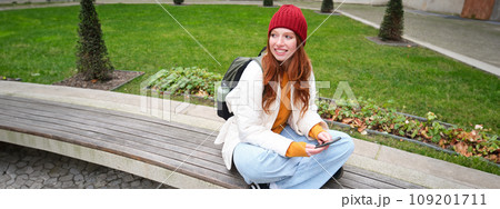 Portrait of stylish young woman, 25 years, sits on bench in park and uses mobile phone, reads online news, messages or watches video on smartphone app, connects to public wifi 109201711