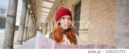 Adventurous redhead girl walks in town with paper map, explores city as tourist, looks for popular tourism attractions, looks around excited and smiles 109201755