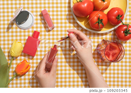 Advertising scene for tomato-based cosmetics. Lipstick, perfume bottle, mirrors and fresh tomatoes are decorated on a checkered background. The woman's hand is holding a tube of tomato red lipstick Advertising scene for tomato-based cosmetics. Lipstick, perfume bottle, mirrors and fresh tomatoes are decorated on a checkered background. The woman's hand is holding a tube of tomato red lipstick 109203345
