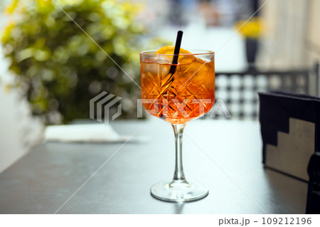 Aperol Spritz Cocktail. Alcoholic beverage based on table with ice cubes and oranges outdoors. Served cocktail with orange slice and straw placed on the table of sidewalk cafe in Italy 109212196