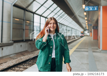 Young worried redhead woman waiting train on station platform, she lost or late, unhappy girl talking on smart phone. Railroad transport concept, Communication banner 109216173