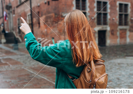 Casual dressed woman 30 years old exploring city streets during international excursion in town. Redhead girl with backpack pointing finger on city street in Gdansk. Traveling Europe in autumn. 109216178