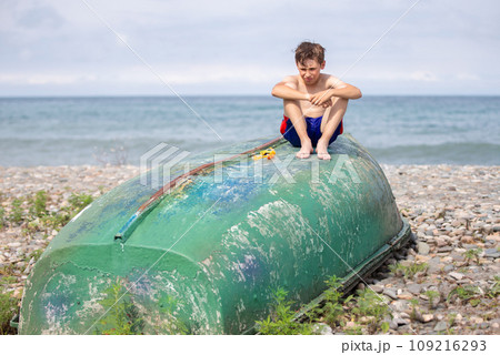 A boy on the beach sits on an old boat washed ashore by a storm. A boy on the beach sits on an old boat washed ashore by a storm. 109216293