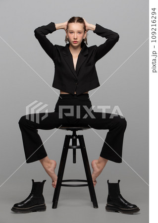 A slender beautiful teenage girl in a black suit sits on a chair barefoot, her shoes are taken off and lie next to her. A slender beautiful teenage girl in a black suit sits on a chair barefoot, her shoes are taken off and lie next to her. 109216294