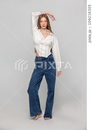 A girl with long white hair in a white shirt and jeans in full length on a gray background. 109216305