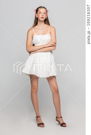 Beautiful romantic young girl in a white short dress posing in the studio on a gray background. Beautiful romantic young girl in a white short dress posing in the studio on a gray background. 109216307