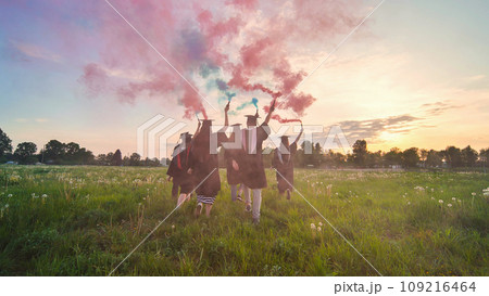 Students graduate with colored smoke walking through the meadow in the evening. 109216464
