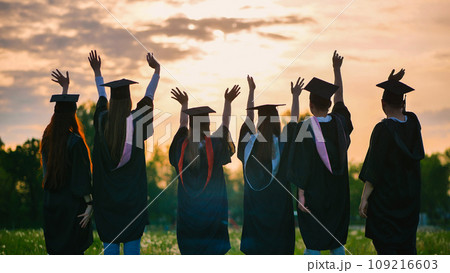 Silhouettes of graduates in black robes waving their arms against the evening sunset. Silhouettes of graduates in black robes waving their arms against the evening sunset. 109216603