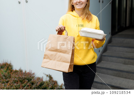 Girl with bag in hand posing on city background. A Female delivery hand with craft shopping bag and box outdoors. Fast Food delivery service, area for your logo or design. Packaging template mock up. 109218534