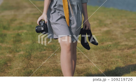 A barefoot girl photographer walks through a meadow with a camera and shoes. A barefoot girl photographer walks through a meadow with a camera and shoes. 109219720