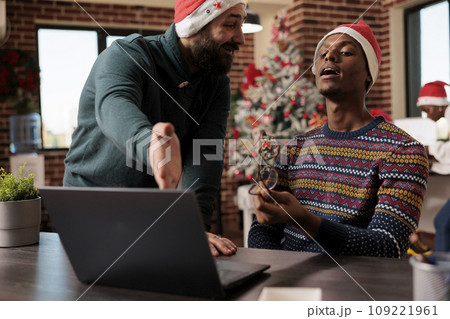 Company team lead in santa hat helping employee with project while working in christmas decorated office. Manager pointing on laptop and explaining task to coworker in festive workplace 109221961