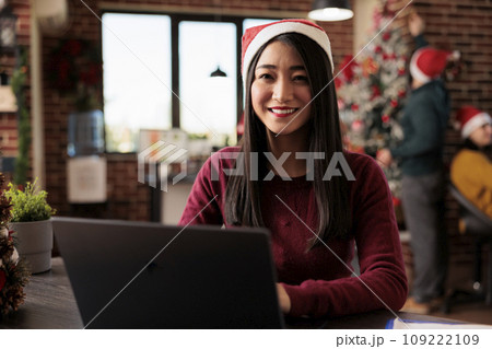 Smiling asian woman worker in santa hat working on laptop in festive workspace portrait. Cheerful corporate employee using computer at desktop in winter holiday decorated office Smiling asian woman worker in santa hat working on laptop in festive workspace portrait. Cheerful corporate employee using computer at desktop in winter holiday decorated office 109222109