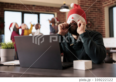 Frustrated tired employee putting santa hat on eyes while working overtime during christmas season in office. Startup company worker managing tasks workload during winter holiday Frustrated tired employee putting santa hat on eyes while working overtime during christmas season in office. Startup company worker managing tasks workload during winter holiday 109222122