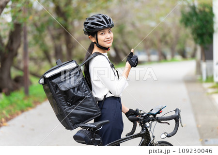 ロードバイクを前にしてデリバリーバックを背負って親指立てているフードデリバリー配達員の女性 ロードバイクを前にしてデリバリーバックを背負って親指立てているフードデリバリー配達員の女性 109230046