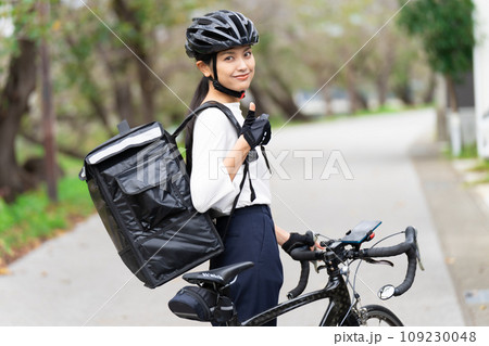 ロードバイクを前にしてデリバリーバックを背負って親指立てているフードデリバリー配達員の女性 ロードバイクを前にしてデリバリーバックを背負って親指立てているフードデリバリー配達員の女性 109230048