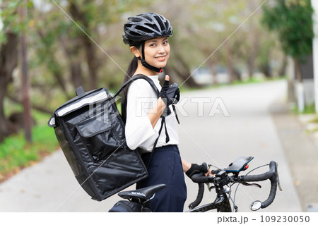 ロードバイクを前にしてデリバリーバックを背負って親指立てているフードデリバリー配達員の女性  109230050