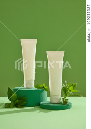 On a green background, two white plastic tubes placed on green podiums with spearmint leaves. Front view for advertising, blank space for design. Spearmint leaves firm the skin and soften dry skin 109231387