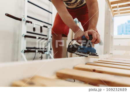 unrecognizable man rubs wooden child bed with power sander or grinder removing old paint 109232689
