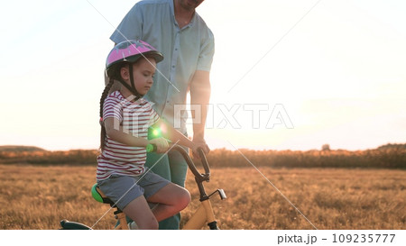 Diligent little girl in helmet rides bicycle with caring father in summer field Diligent little girl in helmet rides bicycle with caring father in summer field 109235777