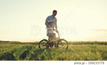 Smiling father teaches preschooler daughter to ride bicycle in summer field Smiling father teaches preschooler daughter to ride bicycle in summer field 109235778