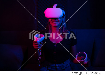Young asian woman using at home Virtual Reality headset with joysticks playing VR video games, neon pink blue light Young asian woman using at home Virtual Reality headset with joysticks playing VR video games, neon pink blue light 109238926