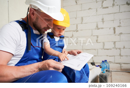 Male builder and child studying building plan in apartment with brick wall on background. Kid in construction helmet sitting on father lap and pointing at architectural drawings. Male builder and child studying building plan in apartment with brick wall on background. Kid in construction helmet sitting on father lap and pointing at architectural drawings. 109240166
