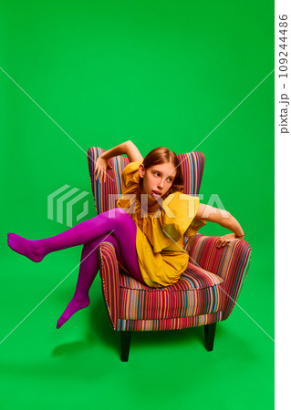 Young attractive girl in unusual, strange, variegated colored outfit and posing extraordinary in retro chair against green background. 109244486