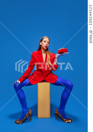 Fashion standards. Young lady posing in unusual, freaky, bright outfit, wearing blue tights, red jacket and animal printed heels holding birthday cake. 109244525