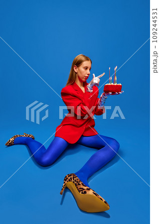 Young lady dressed unusual, freaky, bright outfit, wearing blue tights, red jacket and animal printed heels holding birthday cake and count candles. Young lady dressed unusual, freaky, bright outfit, wearing blue tights, red jacket and animal printed heels holding birthday cake and count candles. 109244531