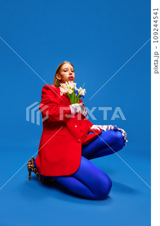 Weird girl in blue tights posing with flowers in unusual, strange, red jacket sitting against blue color studio background. 109244541