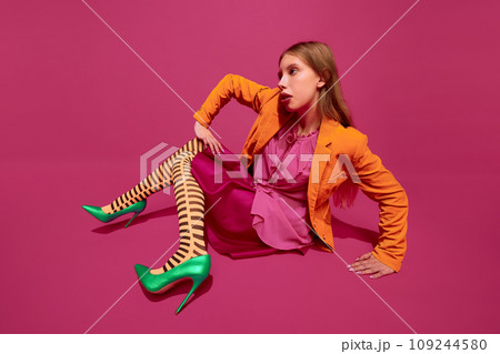 Young girl dressed in stylish colorful striped orange tights, skirt looks like freak, poses sitting against magenta color background background. Young girl dressed in stylish colorful striped orange tights, skirt looks like freak, poses sitting against magenta color background background. 109244580