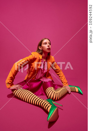Full-length portrait of expressive young girl, lady in unusual, variegated orange striped tights, long skirt and short jacket against magenta background. Full-length portrait of expressive young girl, lady in unusual, variegated orange striped tights, long skirt and short jacket against magenta background. 109244592