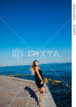 Beautiful woman in black summer dress on a walk by the sea Beautiful woman in black summer dress on a walk by the sea 109245300