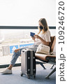 A young teenage girl is sitting at the airport waiting for her flight next to her suitcase. Girl holding a passport in her hands and wearing a mask A young teenage girl is sitting at the airport waiting for her flight next to her suitcase. Girl holding a passport in her hands and wearing a mask 109246720
