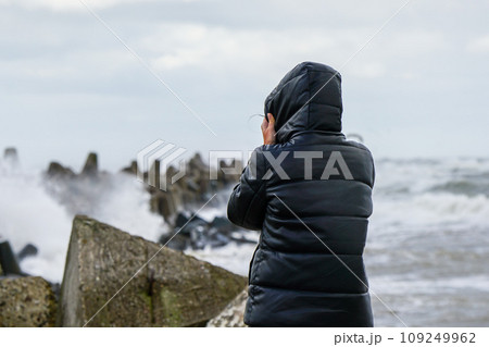 A woman in a black jacket with a hood holds the hood in a strong wind and looks at the stormy sea A woman in a black jacket with a hood holds the hood in a strong wind and looks at the stormy sea 109249962