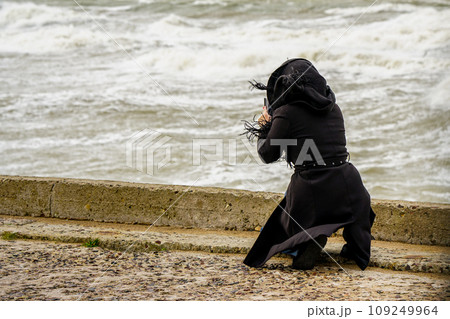 A woman in a black coat with a hood on her knees photographing a stormy sea with her phone A woman in a black coat with a hood on her knees photographing a stormy sea with her phone 109249964