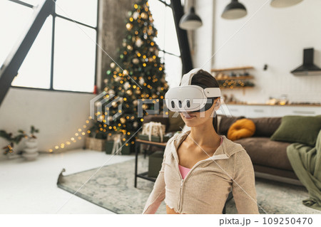 Demonstrating yoga poses, a young woman uses virtual reality glasses near a Christmas tree. 109250470