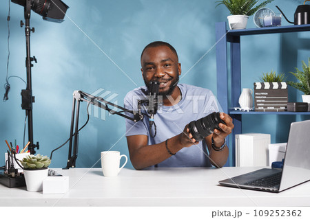 Smiling social media influencer live streaming camera lens review and speaking in microphone. Vlogger showing photography professional equipment and recording video for blog subscribers Smiling social media influencer live streaming camera lens review and speaking in microphone. Vlogger showing photography professional equipment and recording video for blog subscribers 109252362