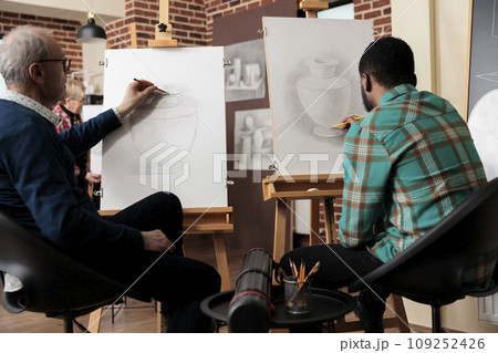 Rear view of two diverse men students sitting at easels practicing still life drawing in art studio. Friends learning new things together, people learn how to draw with pencil during creative workshop 109252426