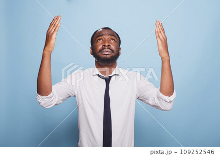 Worried black businessman is praying and raising his hands in a humble gesture towards the sky. African american employee wearing a black tie and a white shirt begs for forgiveness Worried black businessman is praying and raising his hands in a humble gesture towards the sky. African american employee wearing a black tie and a white shirt begs for forgiveness 109252546