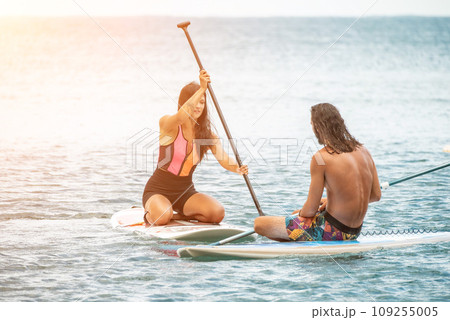 Sea woman and man on sup. Silhouette of happy young woman and man, surfing on SUP board, confident paddling through water surface. Idyllic sunset. Active lifestyle at sea or river. 109255005