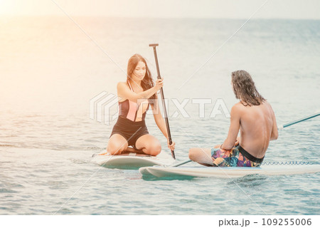 Sea woman and man on sup. Silhouette of happy young woman and man, surfing on SUP board, confident paddling through water surface. Idyllic sunset. Active lifestyle at sea or river. 109255006
