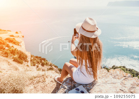 Digital nomad, woman in the hat, a business woman with a laptop sits on the rocks by the sea during sunset, makes a business transaction online from a distance. Freelance, remote work on vacation. Digital nomad, woman in the hat, a business woman with a laptop sits on the rocks by the sea during sunset, makes a business transaction online from a distance. Freelance, remote work on vacation. 109255075