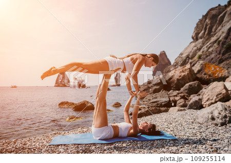 Woman sea yoga. Two happy women practicing yoga on the beach with ocean and rock mountains. Motivation and inspirational fit and exercising. Healthy lifestyle outdoors in nature, fitness concept. 109255114