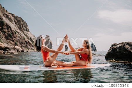 Woman sup yoga. Two happy sporty women practising yoga pilates on paddle sup surfboard. Female stretching doing workout on sea water. Modern individual female hipster outdoor summer sport activity. Woman sup yoga. Two happy sporty women practising yoga pilates on paddle sup surfboard. Female stretching doing workout on sea water. Modern individual female hipster outdoor summer sport activity. 109255548