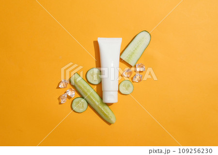 Over orange background, a blank label cosmetic tube displayed with cucumber cut in slices and few ice cubes. Container packaging of skin care branding of Cucumber extract Over orange background, a blank label cosmetic tube displayed with cucumber cut in slices and few ice cubes. Container packaging of skin care branding of Cucumber extract 109256230