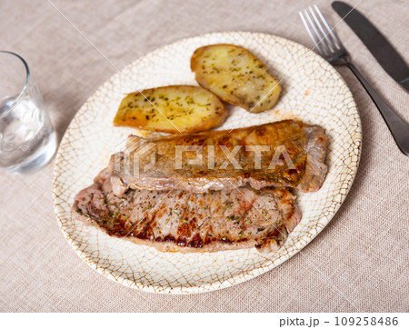 On round white plate lies juicy fried slice of veal tenderloin, garnished with stewed baked potato 109258486